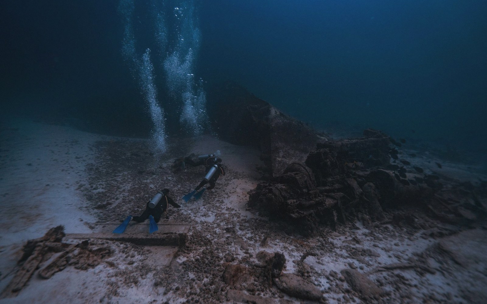 Divers with rising bubble trails examine sunken wreckage resting on the seafloor