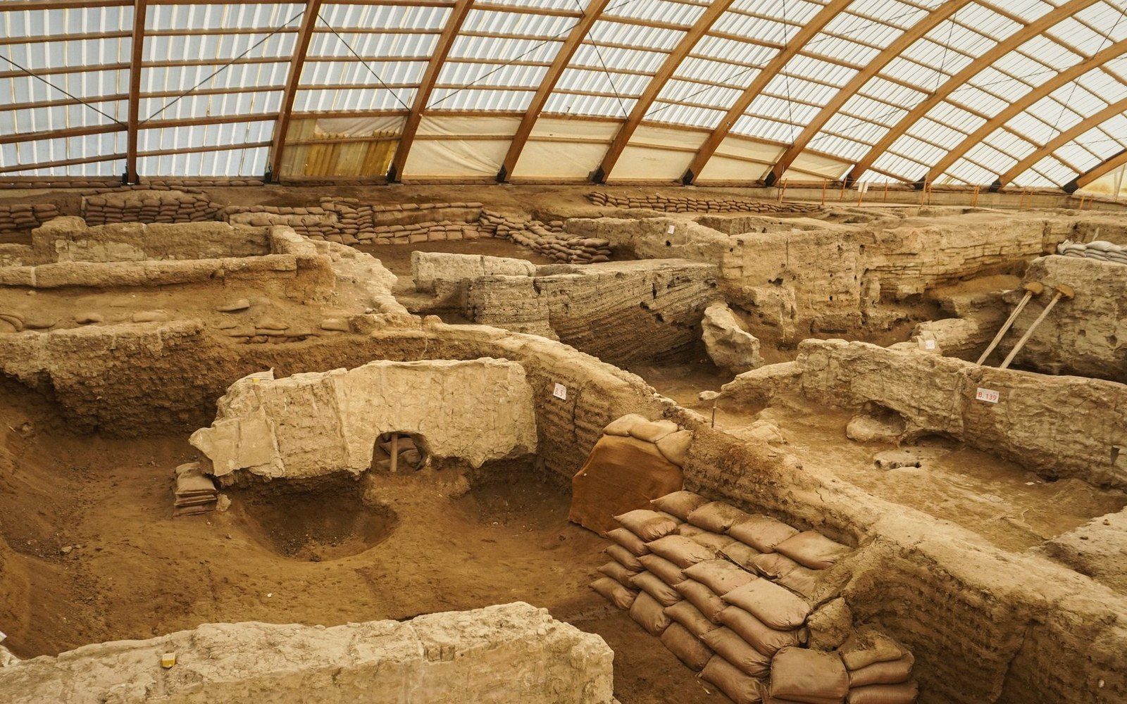 Excavated mudbrick walls and sandbag supports at an ancient settlement site under a protective canopy